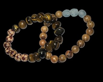 May include: Two beaded bracelets. One bracelet features brown and tan soccer ball-like beads, tiger eye beads, and wooden beads. The other bracelet has wooden beads and light blue beads. The bracelets are on a black background.
