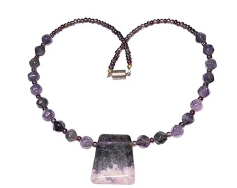 May include: A beaded necklace featuring faceted purple amethyst beads interspersed with small dark metallic spacer beads. A large, trapezoidal pendant with a gradient of dark gray and light pink stone hangs from the center, secured by a metal clasp.
