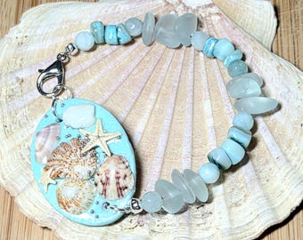 May include: A handmade bracelet featuring a light blue pendant with seashells and starfish, and a strand of light blue and clear beads. The bracelet is set on a large seashell.