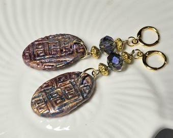 May include: A pair of dangle earrings featuring oval-shaped, textured copper-toned pendants with a raised Greek key pattern. Each earring includes a faceted dark purple crystal bead, gold-toned spacer beads, and a gold-toned lever back closure.