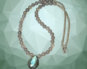 May include: A beaded necklace featuring a strand of gray, round beads. A teardrop-shaped pendant with iridescent blue and green hues hangs from the necklace. The necklace has a silver-colored chain and clasp.
