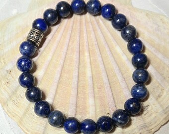 May include: A beaded bracelet featuring dark blue lapis lazuli stones. The bracelet has a silver-colored spacer bead and is displayed on a seashell. The lapis lazuli beads are round and have a polished finish.
