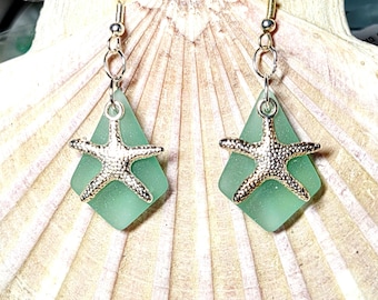May include: A pair of dangle earrings featuring a silver starfish charm over a seafoam green, diamond-shaped glass. The earrings hang from silver-colored hooks and are displayed on a seashell.