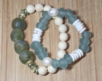 May include: Two beaded bracelets. One bracelet features large, olive-green glass beads and gold-colored spacers. The other bracelet has seafoam-green glass beads, white shell beads, and white pearls. The bracelets are on a light brown wooden surface.