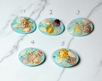 May include: Five oval-shaped, turquoise resin pendants, each adorned with a unique arrangement of seashells, starfish, and small beads. The shells vary in color and size, creating a beachy, nautical theme.