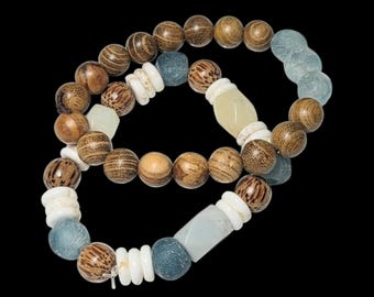 May include: Two beaded bracelets with a variety of natural elements. The bracelets feature wooden beads, white shell accents, and blue-green stone beads. The bracelets are set against a black background.