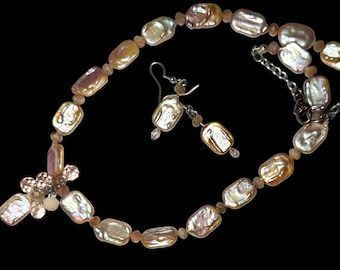 May include: A necklace and earring set featuring iridescent, rectangular pearls in shades of cream, peach, and gold. The necklace has a floral accent and is paired with matching drop earrings. The jewelry is set against a black background.
