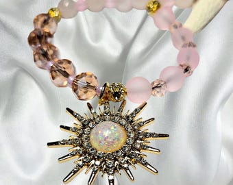 May include: A beaded necklace featuring a sun-shaped pendant with a central iridescent cabochon surrounded by clear rhinestones. The strand consists of alternating light pink matte beads, translucent faceted champagne beads, and small gold-toned accents.