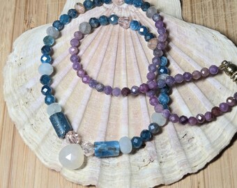 May include: Two beaded necklaces with blue, purple, and clear gemstones rest on a seashell. The necklaces feature various shapes and sizes of beads, including faceted and cylindrical stones. The background is a light brown wooden surface.