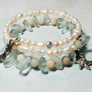 May include: A double-strand bracelet featuring white pearls and light blue sea glass beads. The bracelet has two charms: a mermaid and a starfish.