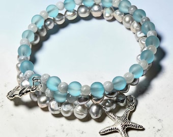 May include: A multi-strand bracelet featuring light blue, white, and gray beads. The bracelet has a silver starfish charm and a silver clasp. The beads are arranged in a spiral pattern, creating a layered effect.