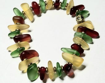 May include: A stretch bracelet featuring irregular, frosted sea glass beads in shades of deep red, forest green, and pale yellow. The beads are interspersed with small, round, metallic gold-toned spacer beads and one larger, textured gold-toned bead.