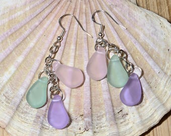 May include: A pair of earrings with silver-colored hooks and chains. Each earring features three teardrop-shaped beads in pastel colors: pink, green, and purple. The earrings are displayed on a large, textured seashell.