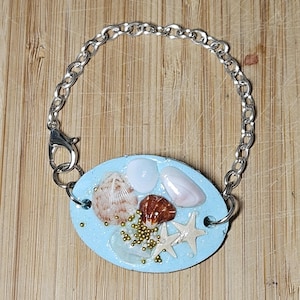 May include: A light blue bracelet with a silver chain and an oval charm. The charm is filled with seashells, a starfish, and gold glitter.