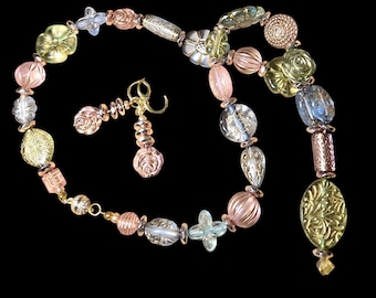 May include: A necklace and earring set featuring a variety of colorful beads. The necklace includes pink, yellow, and clear beads, with a gold chain. The earrings have a gold hook and matching pink beads. The set is displayed against a black background.