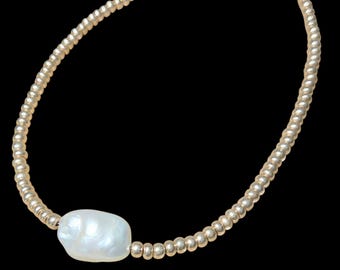May include: A gold beaded necklace with a large, irregular white pearl pendant. The necklace features small, round gold beads strung together, creating a delicate and elegant design. The pearl is centered, adding a focal point to the jewelry.