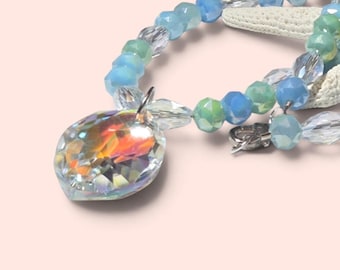 May include: A necklace featuring a large, faceted, iridescent heart-shaped pendant suspended from a strand of alternating light blue, mint green, and clear crystal beads. The beads are multifaceted, reflecting light, and the pendant displays a rainbow-like shimmer.