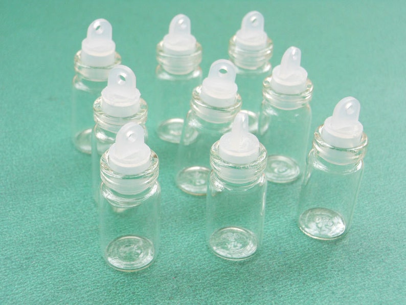 May include: Nine clear glass bottles with white plastic lids and a small loop on top. The bottles are arranged in a semi-circle on a green surface.