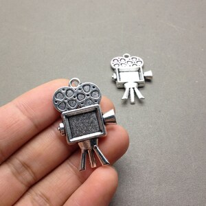 May include: Two silver-toned metal movie camera charms with a film reel design. The charms have a detailed design and are perfect for adding a vintage touch to jewelry or crafts.