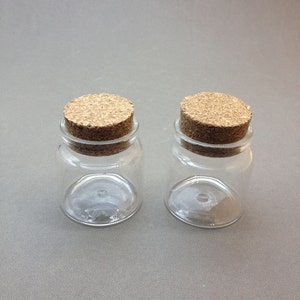 May include: Two small clear glass jars with cork lids. The jars are empty and have a round shape.