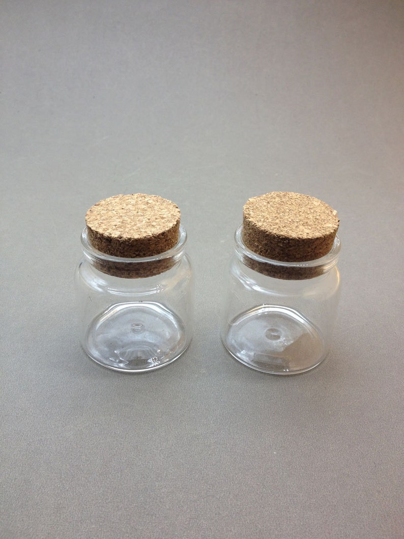 May include: Two small clear glass jars with cork lids. The jars are empty and have a round shape.