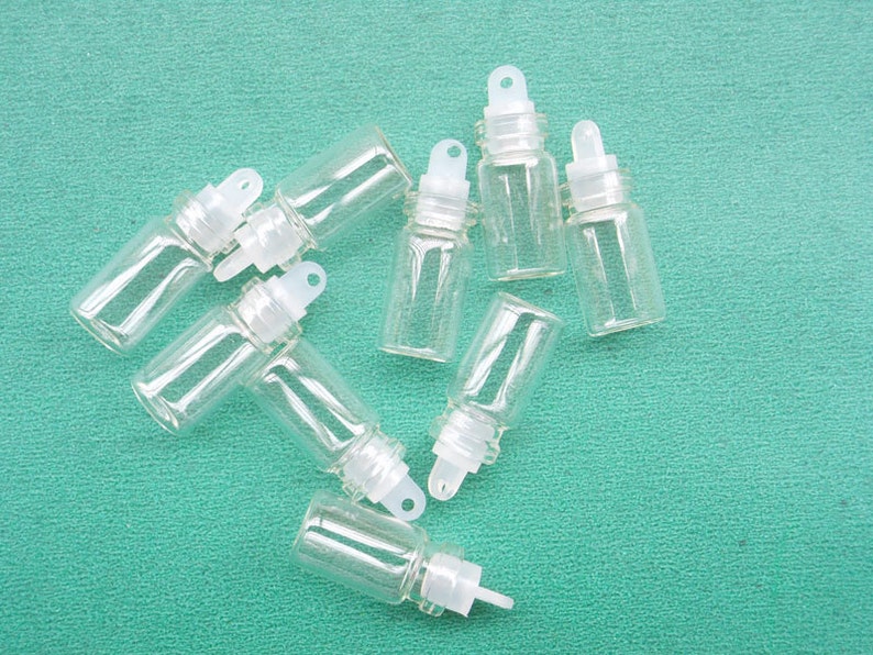 May include: Eight clear glass bottles with white dropper tops and a small loop on the top. The bottles are arranged in a star shape on a green surface.