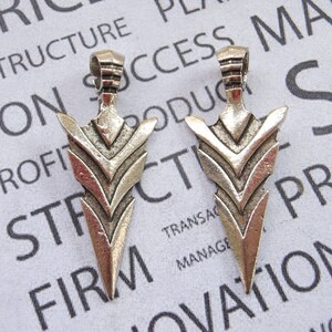 May include: Two silver arrowhead pendants with a geometric design. The pendants are on a white background with text.