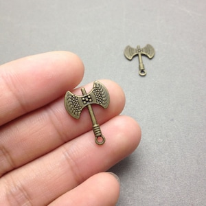 以下が含まれることがあります： 詳細なデザインが施されたアンティークブロンズの斧チャーム2個。斧の頭には装飾的なパターンが施され、ハンドルはねじれています。チャームの上部にはループがあり、ジュエリーやその他のクラフトに取り付けることができます。