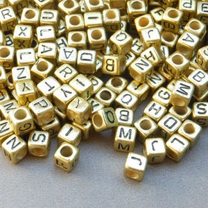 Puede incluir: Un montón de cuentas de letras doradas. Las cuentas son de forma cúbica y tienen letras negras en cada lado.