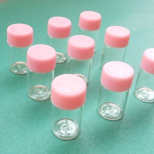 Könnte beinhalten: Neun klare Glasbehälter mit rosafarbenen Deckeln. Die Behälter sind alle gleich groß und gleich geformt und in einem Halbkreis auf einer grünen Oberfläche angeordnet.