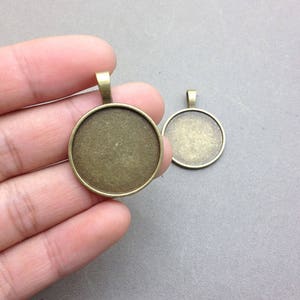 Puede incluir: Dos bandejas redondas de metal de color bronce con un bucle en la parte superior para colgar. La bandeja más grande se sostiene en una mano.
