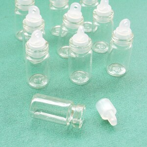 May include: A group of nine clear glass bottles with white plastic lids and a loop on top. The bottles are on a green surface.