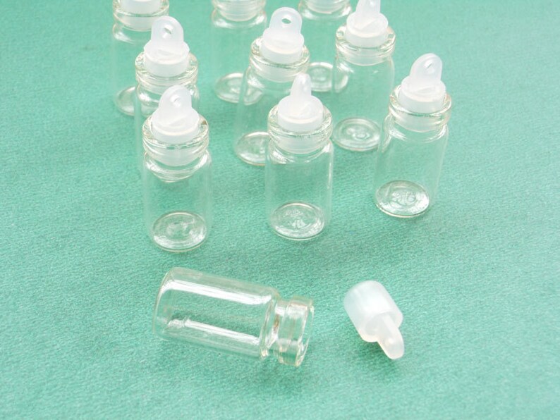 May include: A group of nine clear glass bottles with white plastic lids and a loop on top. The bottles are on a green surface.