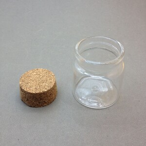 May include: A clear glass jar with a cork lid. The jar is empty and has a round shape.