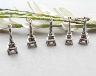 May include: Six silver Eiffel Tower charms, each with a detailed design, are arranged on a white surface with green stems and white flowers in the background.