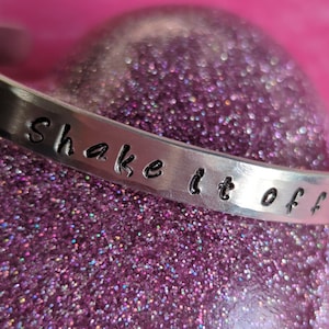 May include: A silver cuff bracelet with the phrase "shake it off" stamped across the front. The bracelet is set against a sparkly, pink background. This piece of jewelry is a stylish accessory.