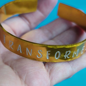May include: A gold-colored metal bracelet with the word "TRANSFORM" engraved in silver lettering.