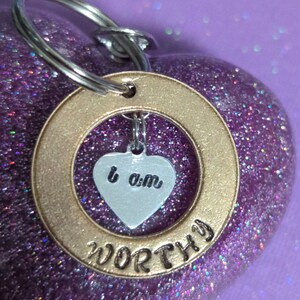May include: A gold-colored keychain with a heart-shaped charm that says "I am" and the word "WORTHY" inscribed around the ring. The keychain is on a purple background with glitter.