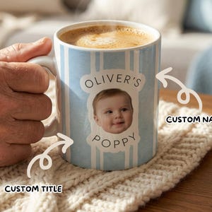 May include: A white ceramic mug with a light blue striped design, filled with coffee. The mug displays a baby's photo and the text "Oliver's Poppy". The mug is held by a person's hand.