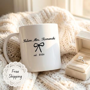 May include: A white ceramic mug with the text "Future Mrs. Romanski" and a bow graphic, alongside a diamond engagement ring in a beige velvet box. The mug also has the text "est. 2026".