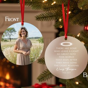 May include: A round Christmas ornament with a red ribbon. The front displays a photo of a woman in a field. The back has text that reads "THERE ARE SOME WHO LEAVE A LIGHT SO GREAT INTO THIS WORLD THAT EVEN AFTER THEY HAVE GONE THE LIGHT REMAINS".