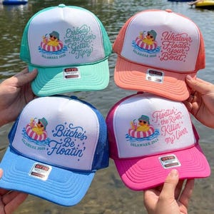 May include: Four trucker hats with white front panels and colored mesh backs. The top left is teal, top right is coral, bottom left is blue, and bottom right is pink. Each hat features a graphic of a rubber duck in a tube and unique text.