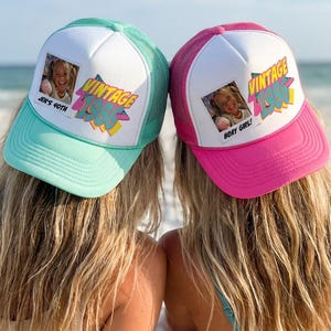 May include: Two trucker hats with white fronts, mesh backs, and curved brims. One is teal, the other pink. Each hat features a photo and the words "VINTAGE 1985" in a colorful design. The teal hat says "Jen's 40th" and the pink hat says "Sexy Girl!"