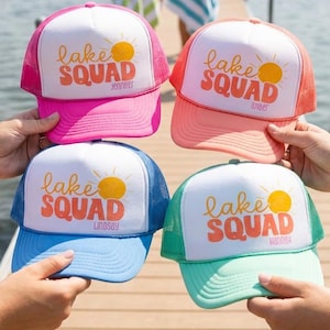 Puede incluir: Cuatro gorras de camionero coloridas con el texto "Lake Squad" y un gr&aacute;fico de sol. Cada gorra tiene un esquema de color diferente: rosa, coral, azul y verde azulado. Las gorras son sostenidas por personas en un muelle de madera cerca de un lago.