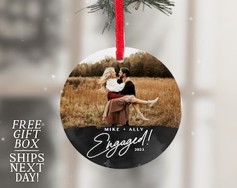 May include: Black and white round Christmas ornament with a photo of a couple in a field. The text "Mike + Ally Engaged! 2023" is written on the ornament.