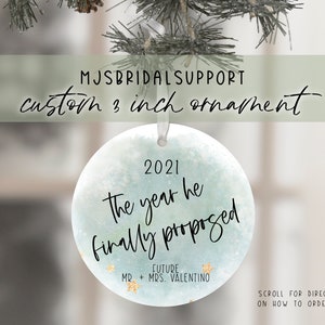 May include: A 3 inch round blue and white watercolor ornament with gold glitter accents. The ornament says "2021 The year he finally proposed" with the text "Future Mr. & Mrs. Valentino" below.