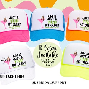May include: A white trucker hat with a mesh back in various colors. The hat features a personalized design with a martini glass and the text "Just a Tini Bit Older" and a space for a photo. The text "13 Colors Available" and "Totally Custom Text" is also visible.