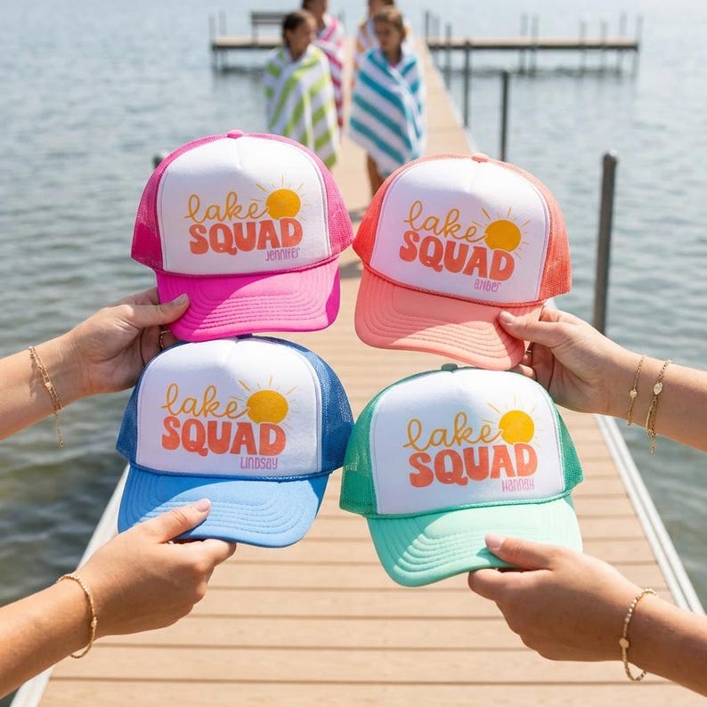 Puede incluir: Cuatro gorras de camionero coloridas con el texto "Lake Squad" y un gr&aacute;fico de sol. Cada gorra tiene un esquema de color diferente: rosa, coral, azul y verde azulado. Las gorras son sostenidas por personas en un muelle de madera cerca de un lago.