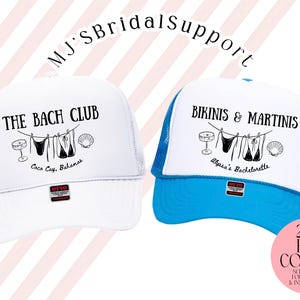 May include: Two white trucker hats with mesh backs. One hat has "THE BACH CLUB" printed on the front, the other reads "BIKINIS & MARTINIS". Both hats feature a beach-themed graphic and text. The hats are for a bachelorette party.