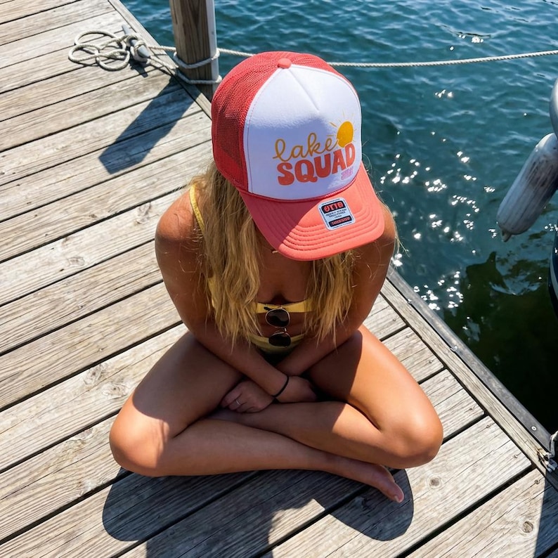 Puede incluir: Gorra de camionero coral y blanca con el texto "Lake Squad" y un gr&aacute;fico de sol amarillo. La gorra tiene una parte trasera de malla y una visera curva. La gorra la lleva una persona sentada en un muelle de madera cerca del agua.